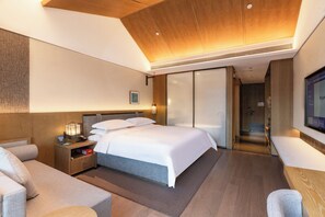 A modern hotel room with a large bed, a wooden headboard, a bedside table with a lamp, a seating area with a sofa, and a television mounted on the wall. - Luoyang Huamao Chunsong Hotel (Luoyang)