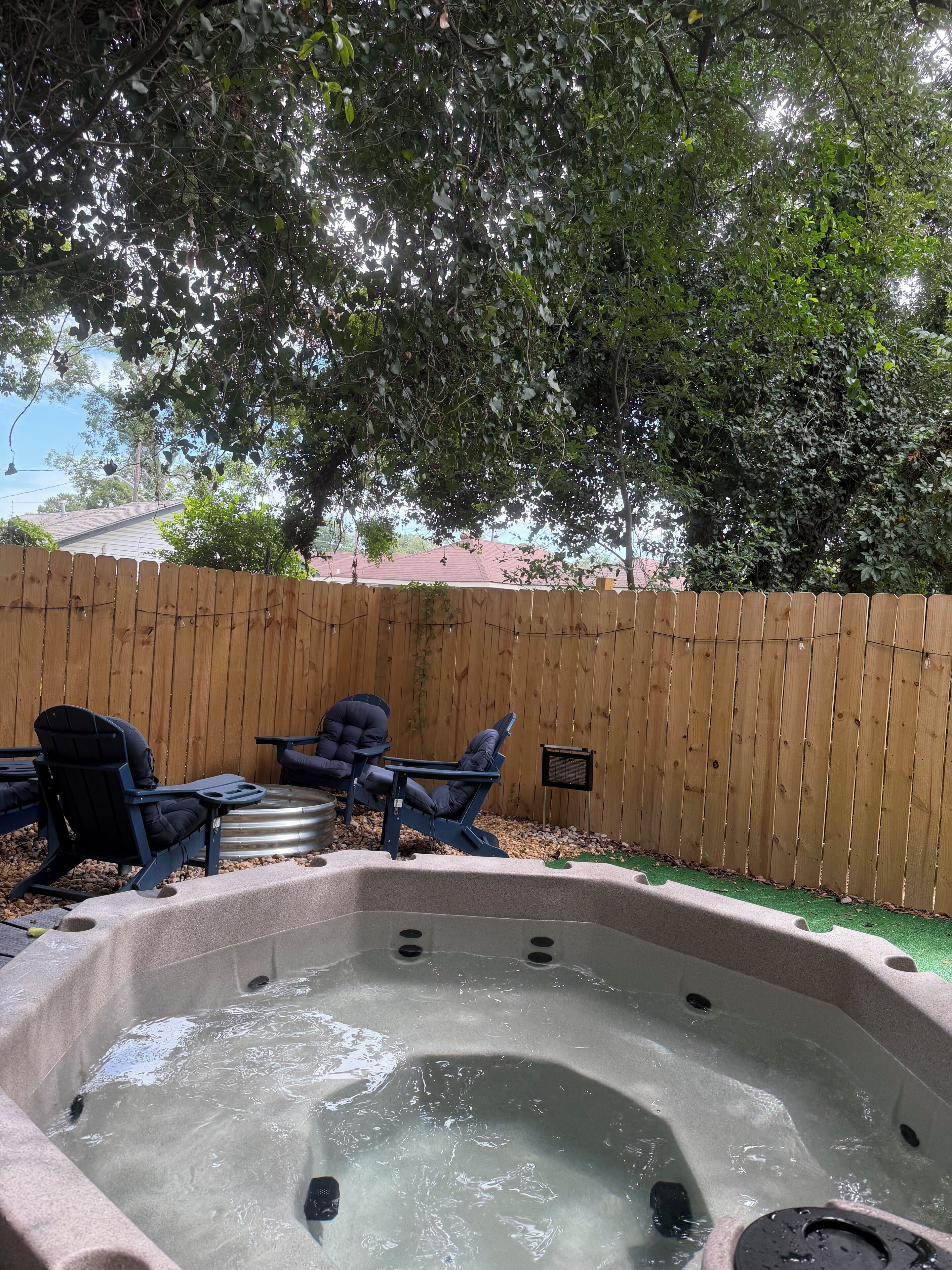 A hot tub with two chairs and a table, surrounded by a wooden fence and trees.