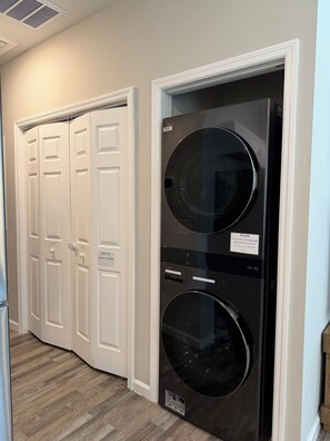 A laundry room with a front-loading washer and dryer, a white door with a 'DO NOT ENTER' sign, and a wooden floor.