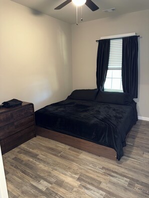 A bedroom with a bed, a dresser, a ceiling fan, a window with curtains, and a wooden floor.