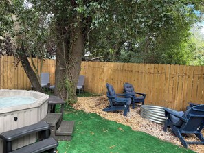 Un patio trasero con un jacuzzi, dos sillas Adirondack, una valla de madera y un árbol.