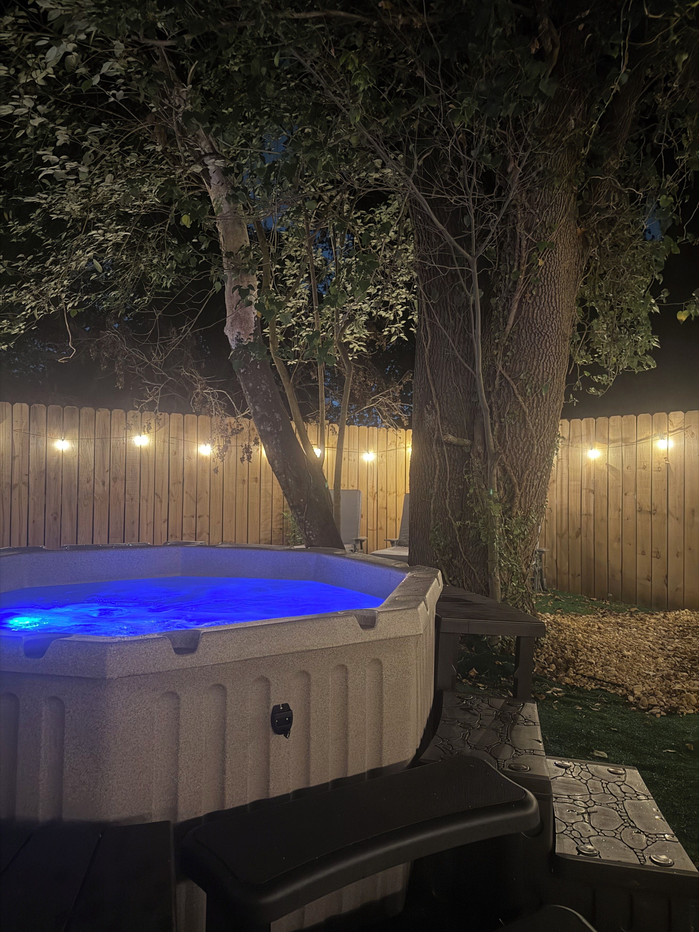 A hot tub with a blue light, surrounded by a wooden fence and trees.