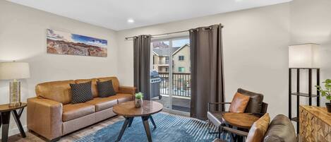 Un salon avec un canapé, une table basse et des fauteuils, un balcon avec des rideaux et un tableau accroché au mur.