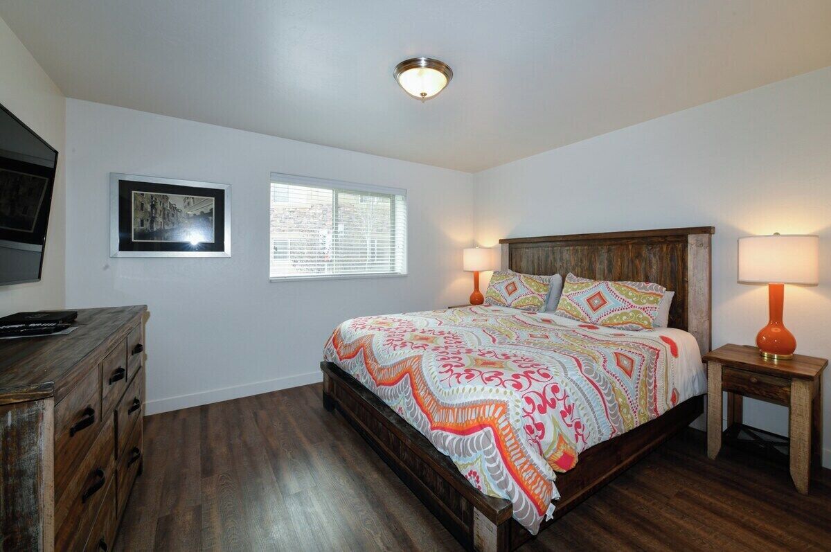 A bedroom with a wooden bed, two bedside lamps, a window with blinds, a dresser, and a framed picture on the wall.