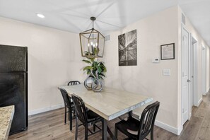 Uma sala de jantar com mesa, cadeiras, luminária pendente, planta e uma geladeira preta.