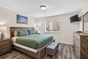 Quarto com cama, mesas de cabeceira, penteadeira, televisão e janela com persianas.