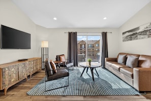 A modern living room with a sofa, a coffee table, a TV, and a view of buildings through large windows. - 4-K Sand Flats Redcliff Condo (Top Floor/Pool Side) (Moab)