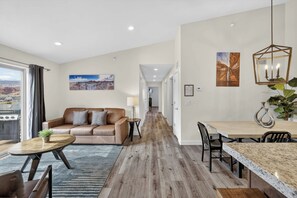 A modern living room with a brown leather sofa, a dining table with chairs, a wooden floor, and a view through a large window. - 4-K Sand Flats Redcliff Condo (Top Floor/Pool Side) (Moab)