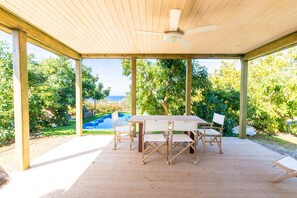 Eine überdachte Terrasse mit Tisch und Stühlen, Holzbalken und Blick auf ein Schwimmbad und Grünflächen.