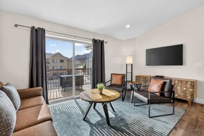 A modern living room with a sofa, armchair, and round table, a TV mounted on the wall, and a balcony with a view of buildings. - 4-L Stargazing Redcliff Condo (Top Floor) (Moab)