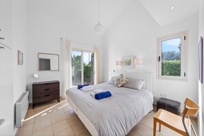 Ein Schlafzimmer mit Bett, Kommode, Stuhl, Schreibtisch und einem Fenster mit Blick auf Grünflächen.