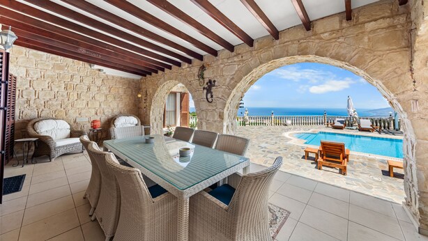 Un patio cubierto con una mesa de vidrio, sillas de mimbre, un muro de piedra y vistas al mar.