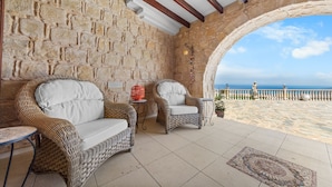 Habitación de paredes de piedra con muebles de mimbre, suelo de baldosas y vista al mar.
