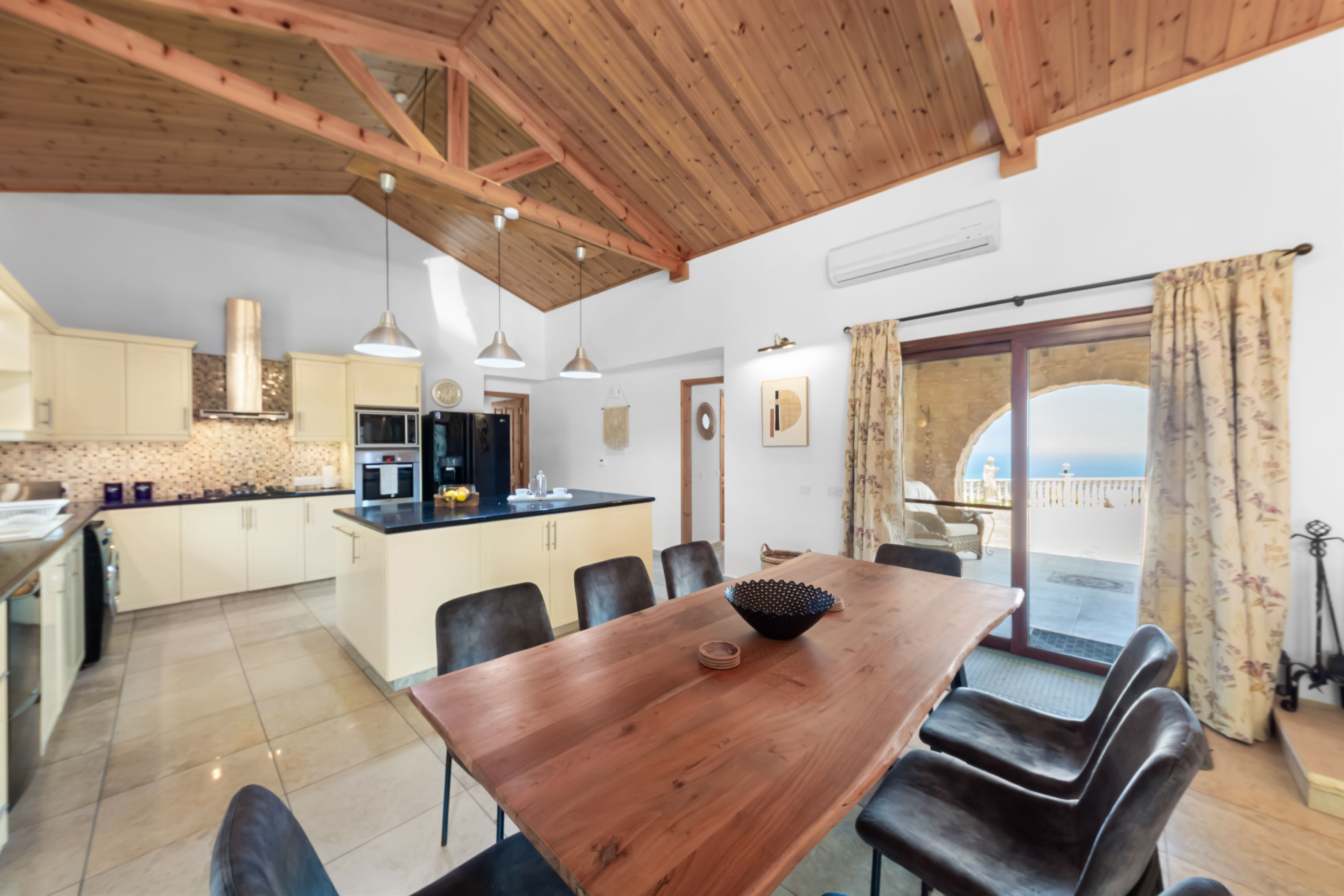 A modern kitchen with wooden beams, a dining area with a wooden table, and a view of the sea through a glass door.
