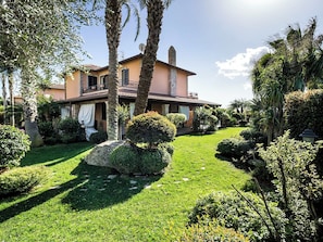 Una casa a due piani con un balcone, immersa in un giardino curato con arbusti e un prato.