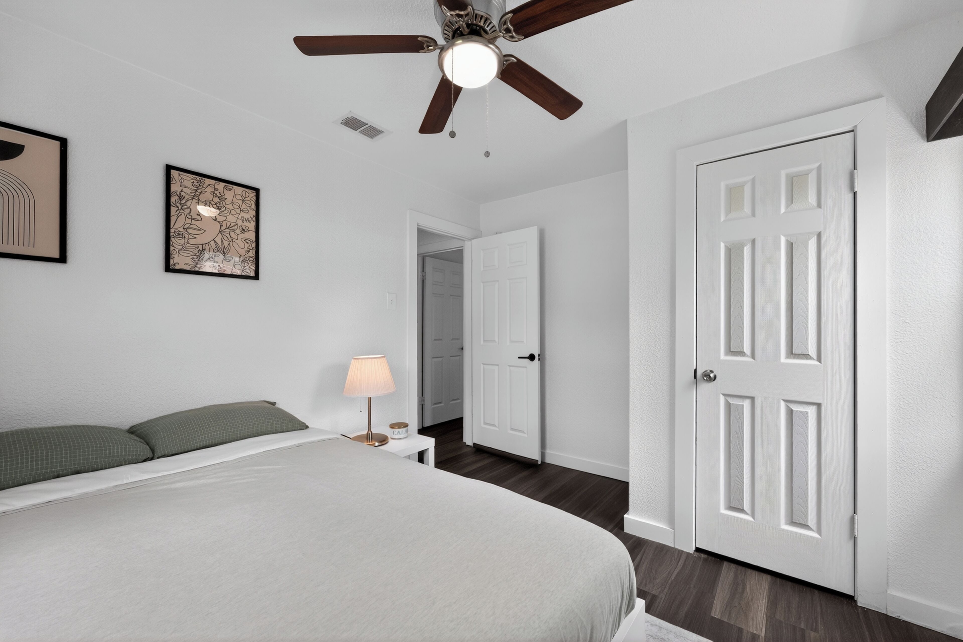 A bedroom with a bed, a ceiling fan, a nightstand with a lamp, and a door.