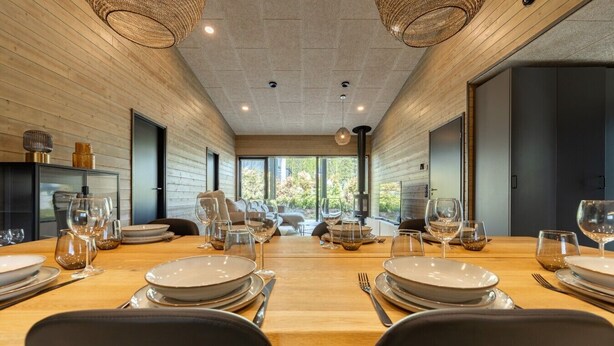 Une salle à manger moderne avec une longue table dressée pour le repas, des murs en bois et de grandes fenêtres.