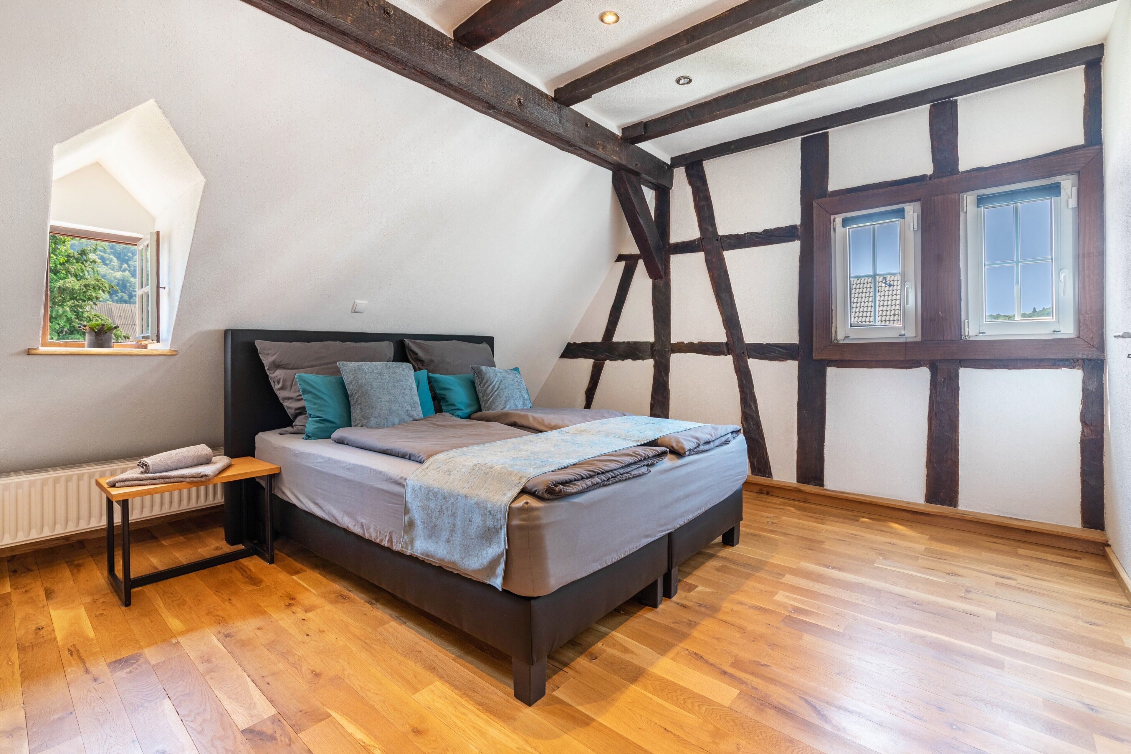 A bedroom with a sloped ceiling, exposed wooden beams, a bed with pillows, a small table, and a window with a view.