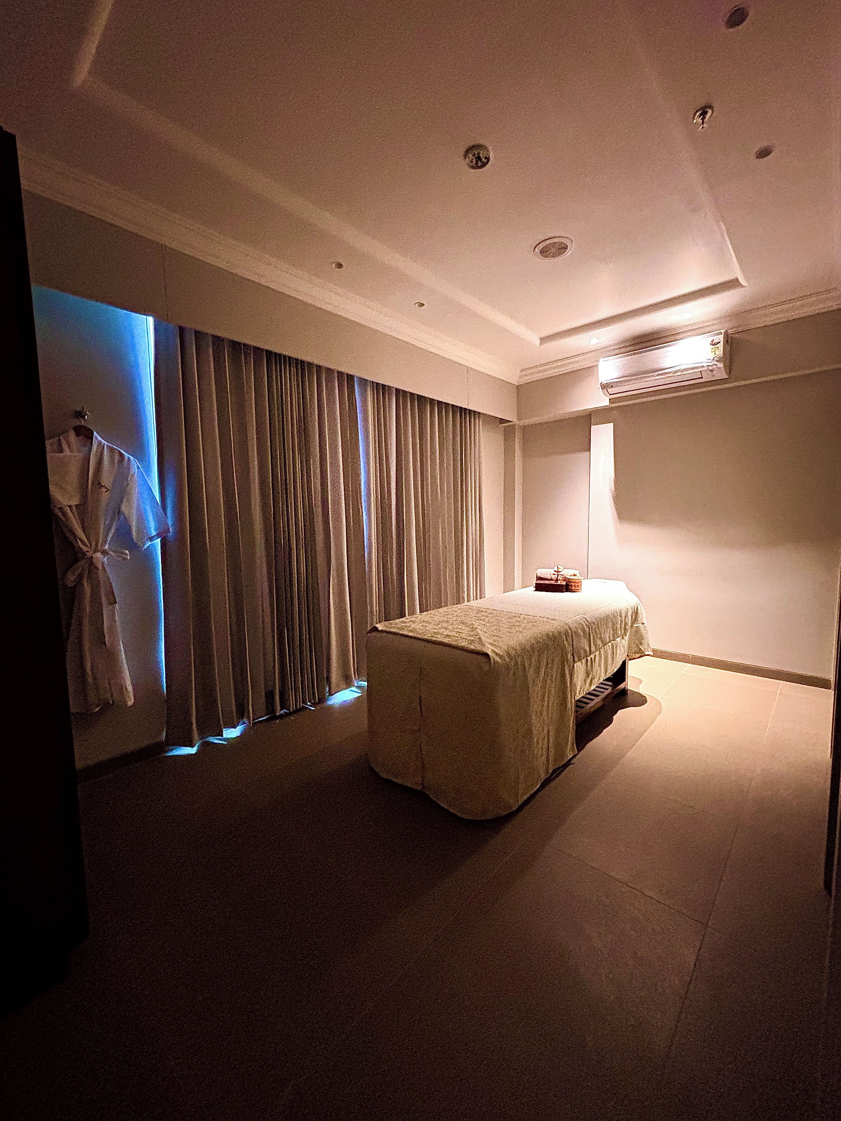 A hotel room with a bed, a window with curtains, an air conditioning unit, and a robe hanging by the window.