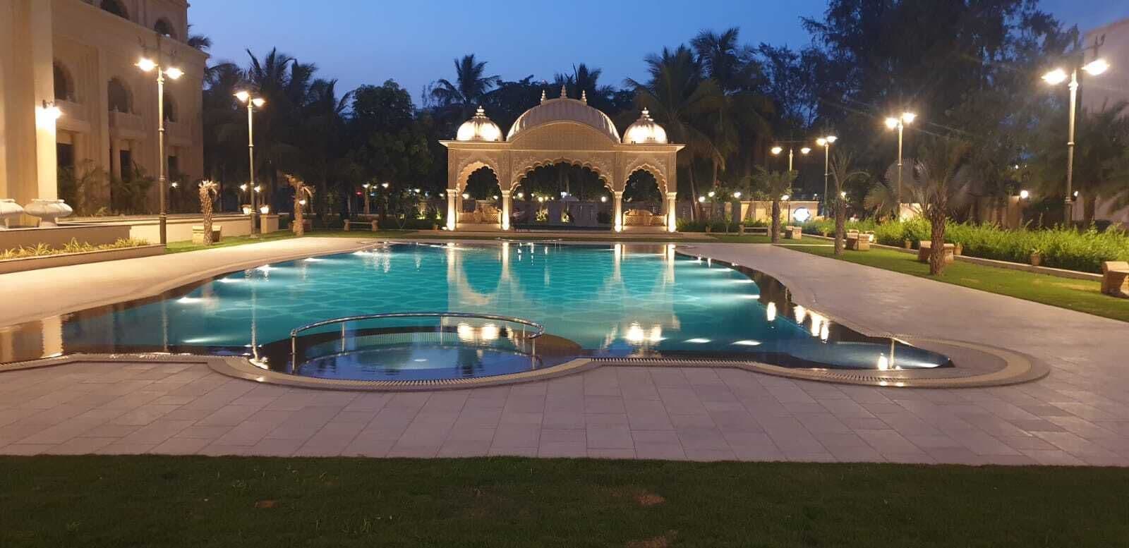 Piscina com fonte, rodeada por um jardim e um edifício com arcos.