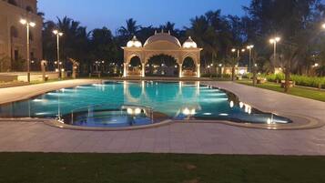Piscina com fonte, rodeada por um jardim e um edifício com arcos.