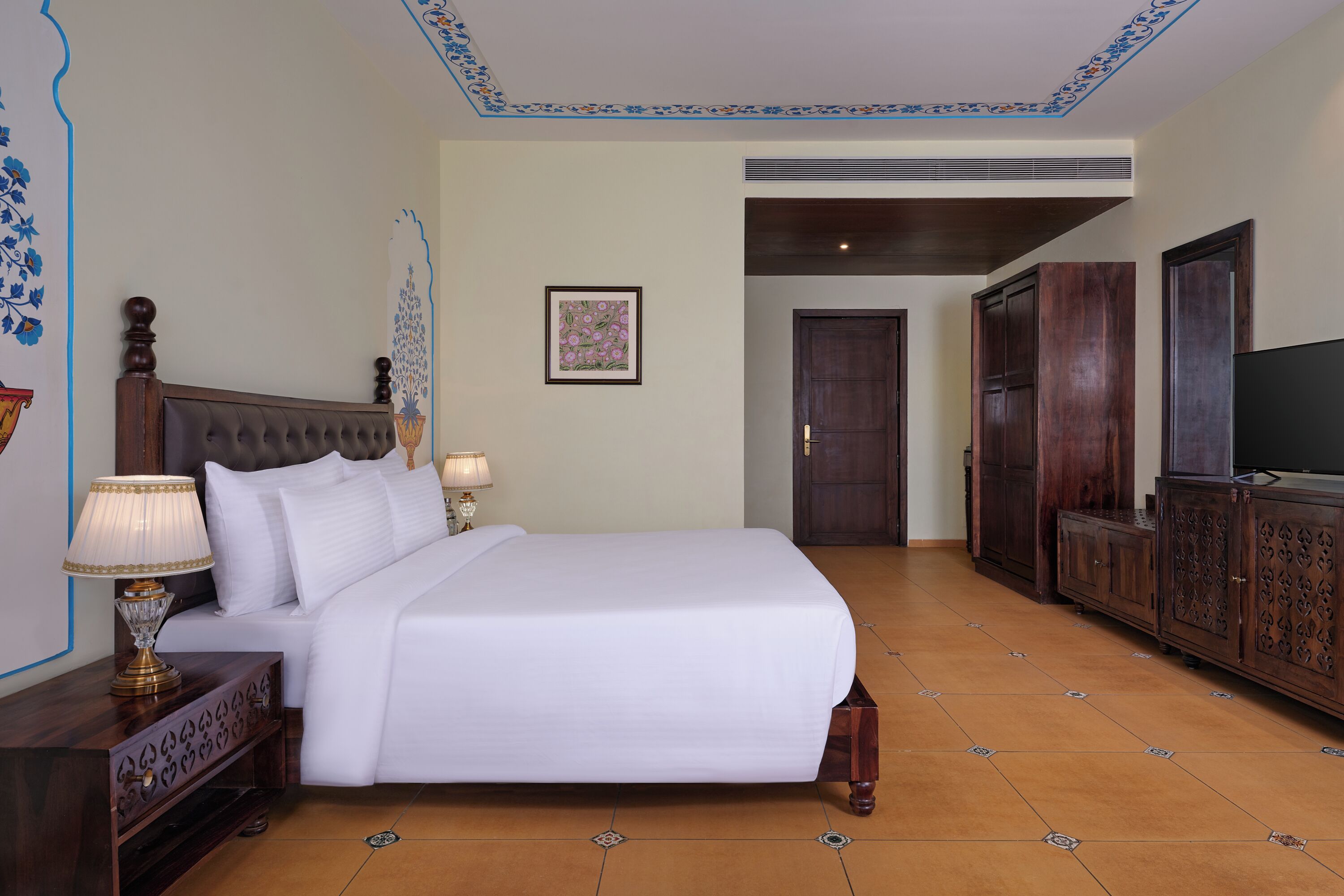 A hotel room with a large bed, wooden headboard, a nightstand with a lamp, a wardrobe, and a television.