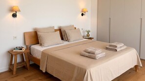 Ein ordentlich gemachtes Bett mit einer beigen Tagesdecke und Kissen, zwei Nachttischlampen, einem kleinen Nachttisch mit Büchern und Gläsern sowie ein gefaltetes Handtuch auf dem Bett.