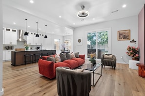 Eine moderne Küche mit weißen Schränken, einer zentralen Kochinsel, Pendelleuchten und einem roten Sofa in einem Wohnbereich mit einem Glastisch und einem grauen Sessel.