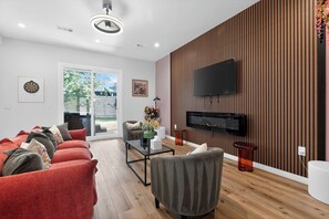Ein modernes Wohnzimmer mit einer Couch, einem Couchtisch, einem an einer Holzwand montierten Fernseher und einer Glastür, die zu einem Außenbereich führt.