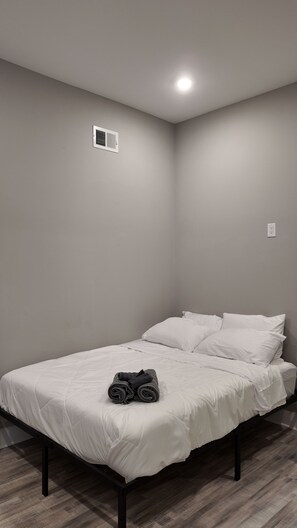 Ein Einzelbett mit weißer Bettwäsche und zusammengerollten Handtüchern, ein Lüftungsgitter an der Wand sowie eine Steckdose.