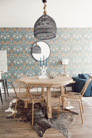 Ein Essbereich mit einem runden Holztisch, Korbstühlen, einem Kuhfellteppich, einer hängenden schwarzen Pendelleuchte und einer Wand mit Blumenpapier.