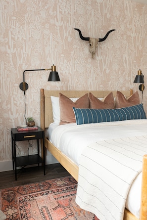 Ein Schlafzimmer mit einem Holzkopfteil, einer Wand mit Muster, einer Totenkopf-Dekoration, zwei Nachttischlampen, einem Nachttisch mit Pflanze und einer Streifen-Decke.