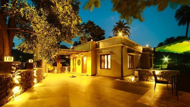 A well-lit building with a paved courtyard, stone walls, and a large tree. - Bungalow Seven (Mount Abu)