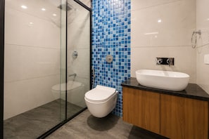 Une salle de bain moderne avec une douche à parois de verre, des toilettes blanches, un mur recouvert de mosaïques bleues et un meuble-lavabo en bois avec un lavabo blanc.