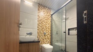 A bathroom with a wooden cabinet, a white sink, a toilet, and a glass shower enclosure. - Bungalow Seven (Mount Abu)