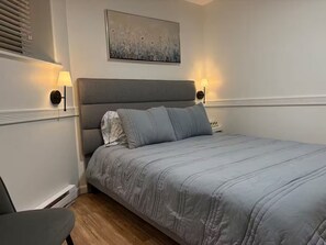 A bedroom with a grey upholstered headboard, a grey bedspread, two bedside lamps, and a painting above the bed. - Amazing 2BR Basement Suite (Langley Township)