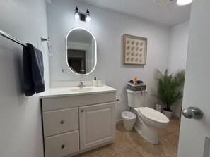 A bathroom with a white vanity, a round mirror with lights, a toilet, a trash can, and a potted plant. - Amazing 2BR Basement Suite (Langley Township)