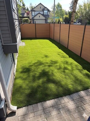 A backyard with artificial green grass, a wooden fence, and a house in the background. - Amazing 2BR Basement Suite (Langley Township)
