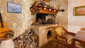 Ein rustikaler Kamin aus Stein mit hölzernem Sims, eine Gitarre, ein Korbstuhl und ein Holzstapel.