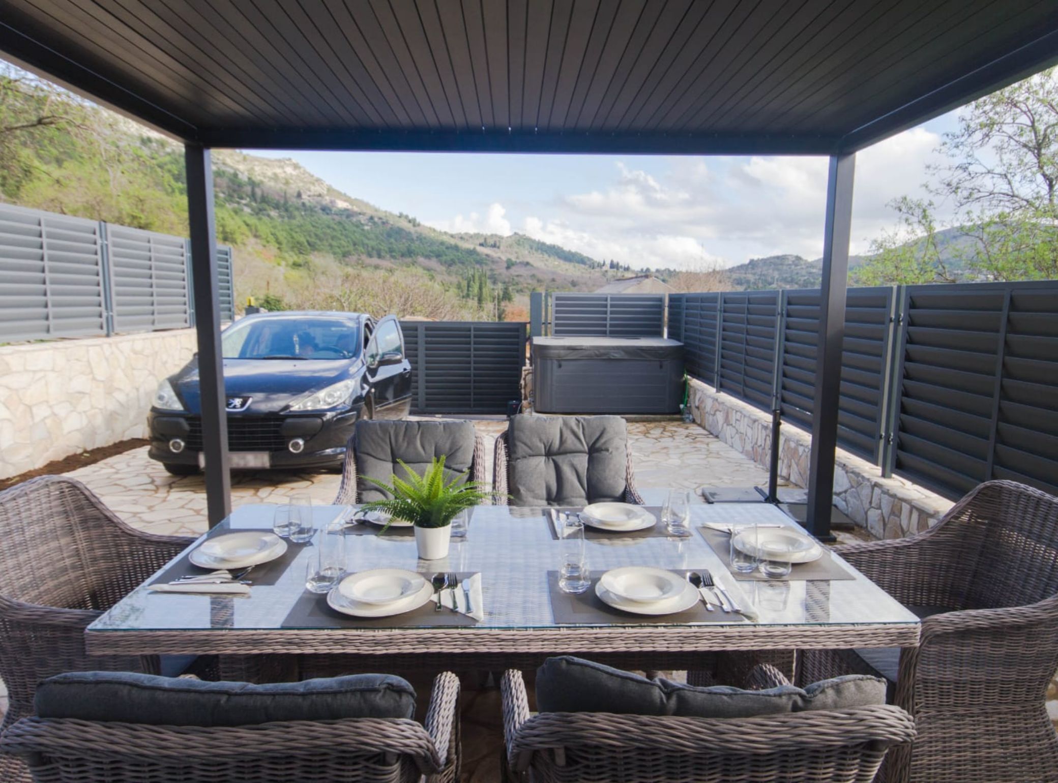 Een terras buiten met een tafel voor vier personen, omringd door rieten stoelen, een auto en een hottub.