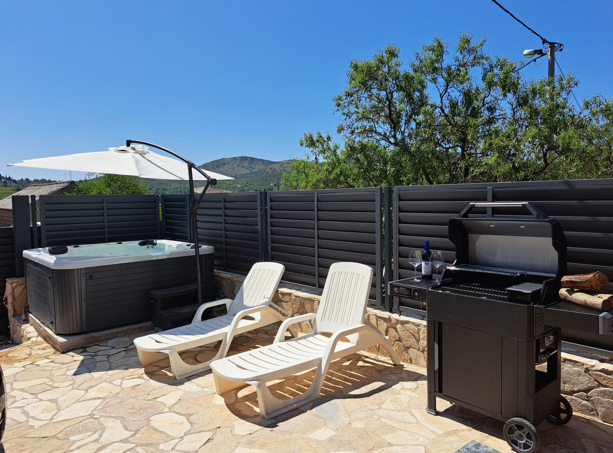 An outdoor patio with a hot tub, lounge chairs, a grill, and a stone-tiled floor.