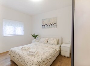 A bedroom with a bed, a nightstand, a window, a wall-mounted painting, and a potted plant.