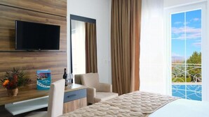 Habitación de hotel con cama, televisor montado en la pared, escritorio con silla, ventana con cortinas y vistas al exterior.