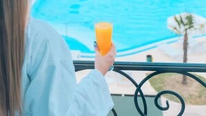 Una persona sosteniendo un vaso de jugo de naranja, mirando una piscina con vistas a árboles y un edificio al fondo.