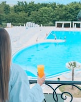 Una persona sosteniendo un vaso de jugo de naranja, mirando una piscina con vistas a árboles y un edificio al fondo.
