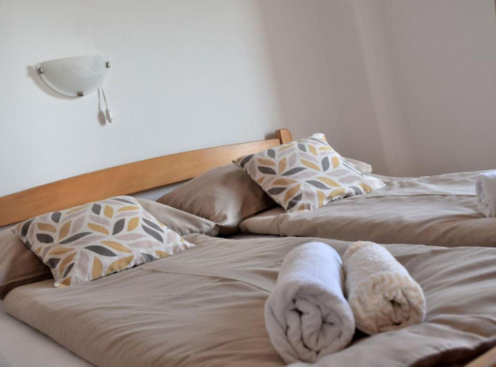 Habitación de hotel con cabecera de madera, dos camas con ropa de cama color beige, almohadas estampadas y una lámpara fijada a la pared.