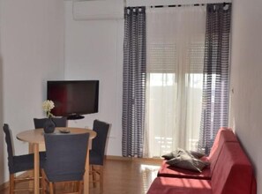 A room with a TV, a dining table with chairs, a red sofa, and a window with curtains. - Andrija Sea View Apartments (Rab)