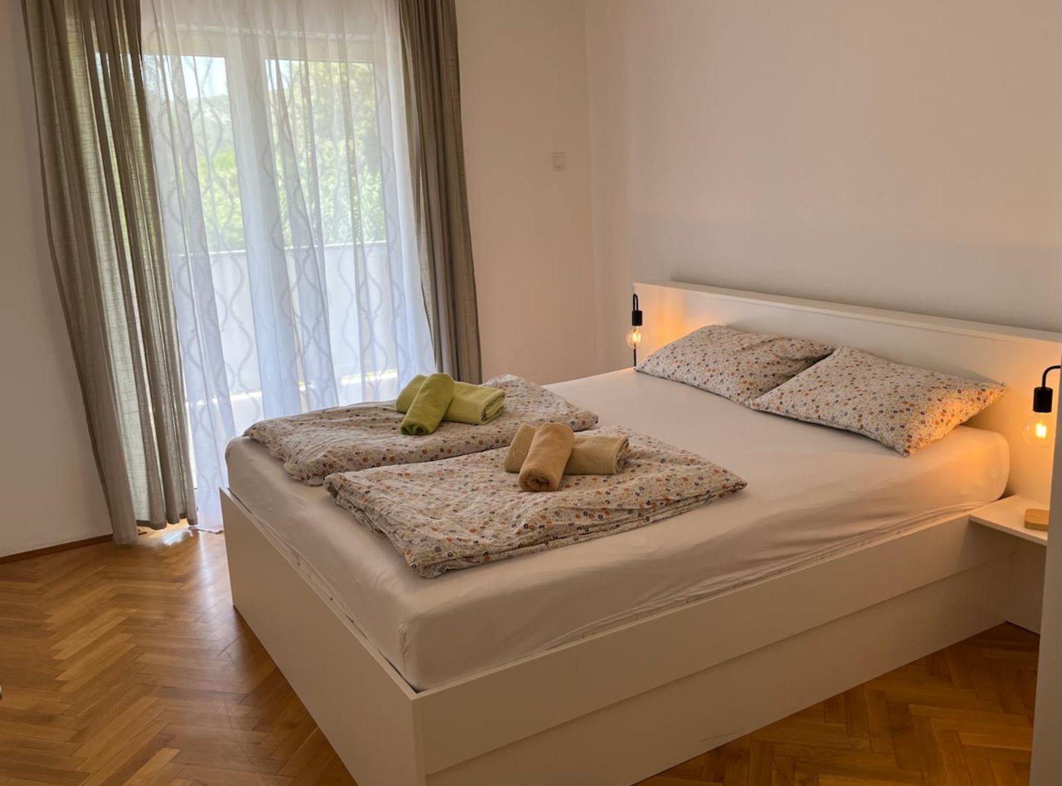 A neatly made bed with floral bedding, a bedside lamp, and a window with sheer curtains.