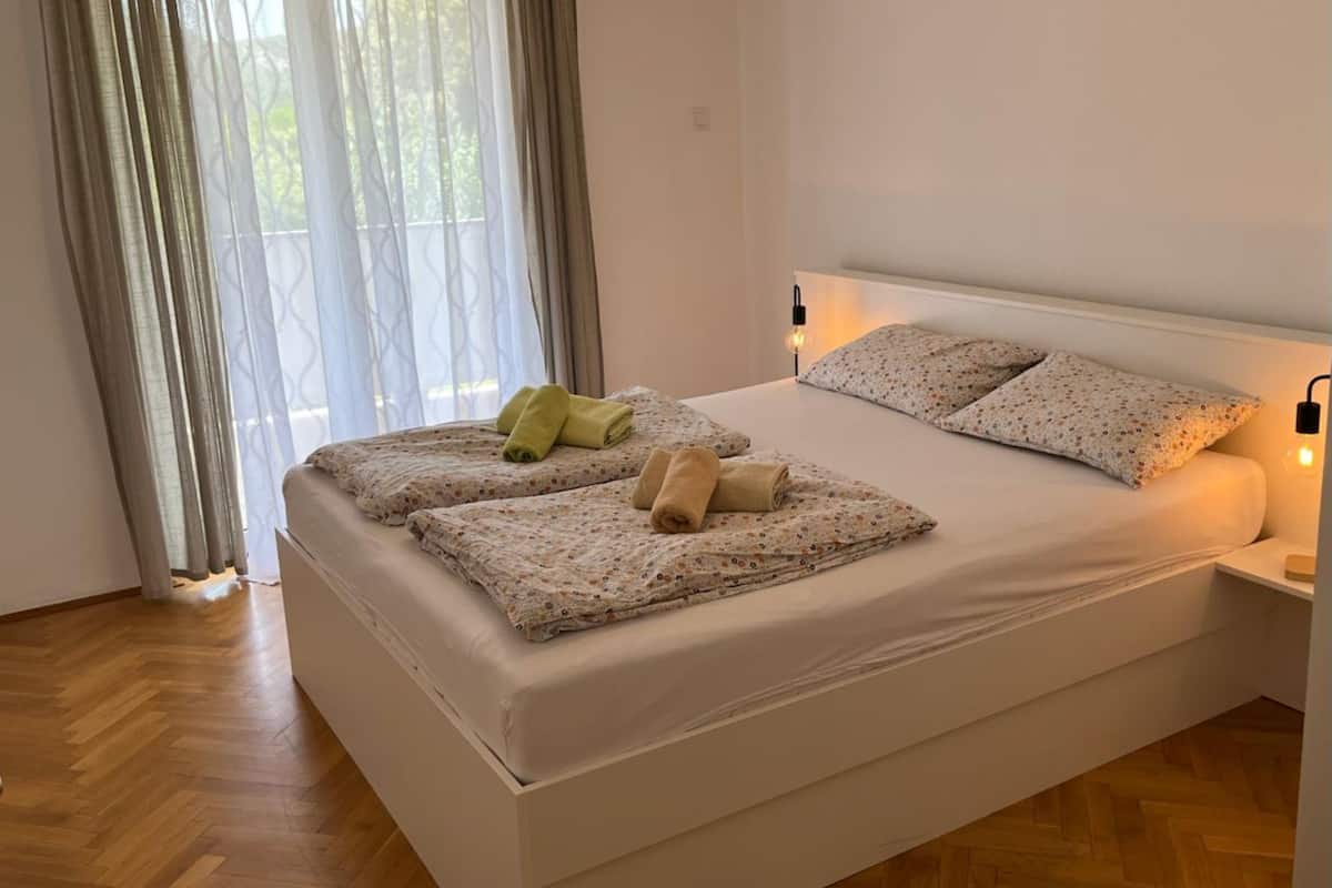 A neatly made bed with floral bedding, a bedside lamp, and a window with sheer curtains.