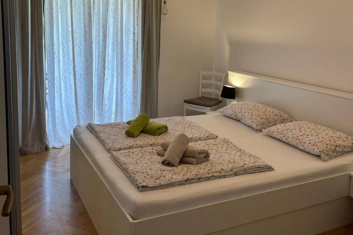 A neatly made bed with floral-patterned bedding, a bedside lamp, and a chair with a cushion.
